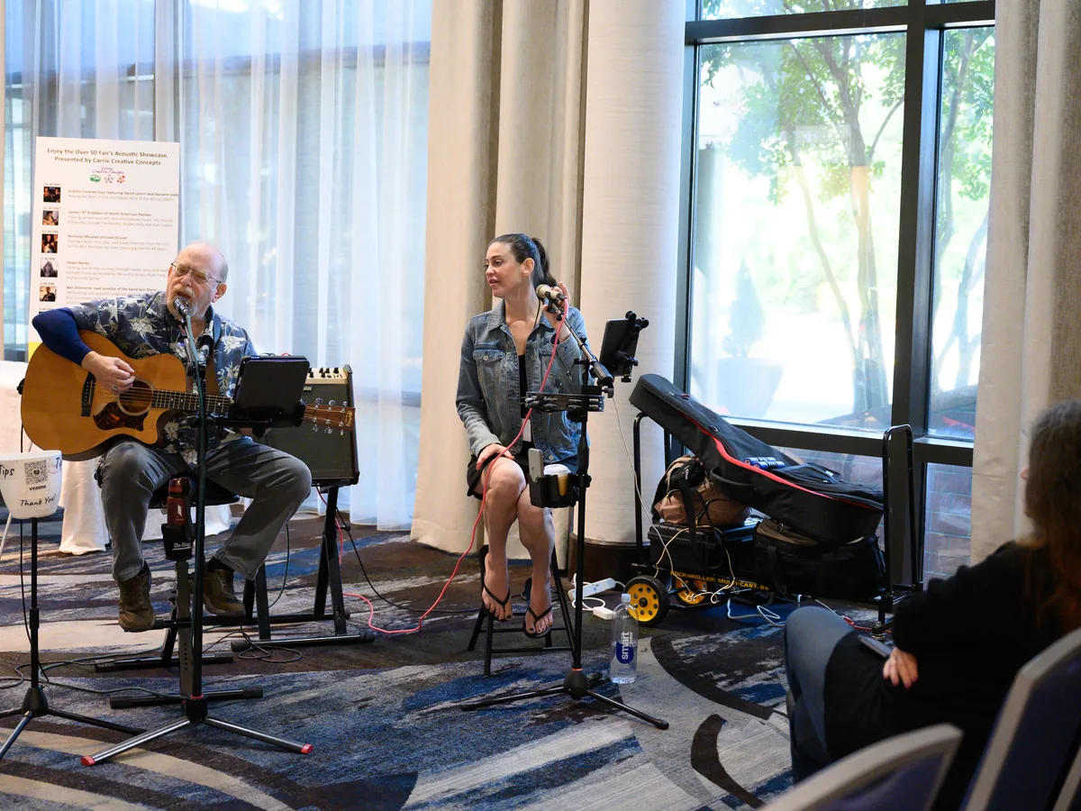 Two musicians performing at the Over 50 Fair, a man playing acoustic guitar and a woman singing into a microphone, beside a window full of daylight