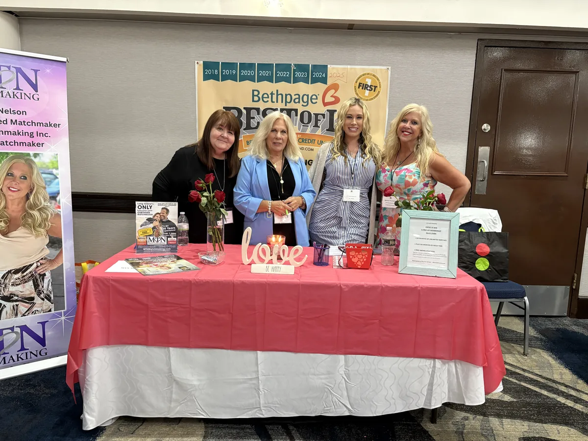 Four women standing behind a table draped in red at the Over 50 Fair, with roses, a candle and a banner of Best of Long Island awards behind them