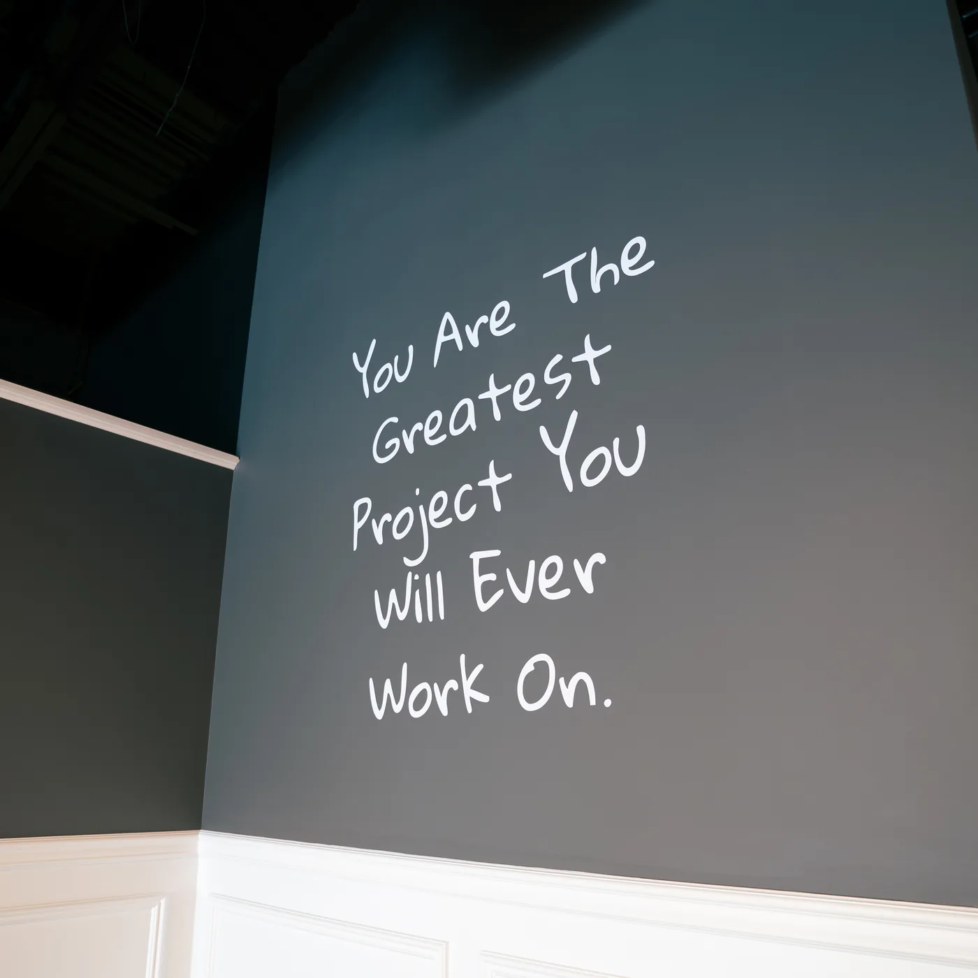 A dark grey wall hand-lettered with the words You Are The Greatest Project You Will Ever Work On.